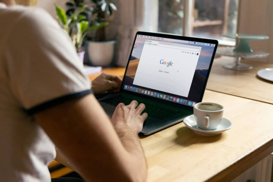 Persona utilizando un portátil con la página principal de Google abierta, mientras disfruta de una taza de café en un ambiente de trabajo informal.