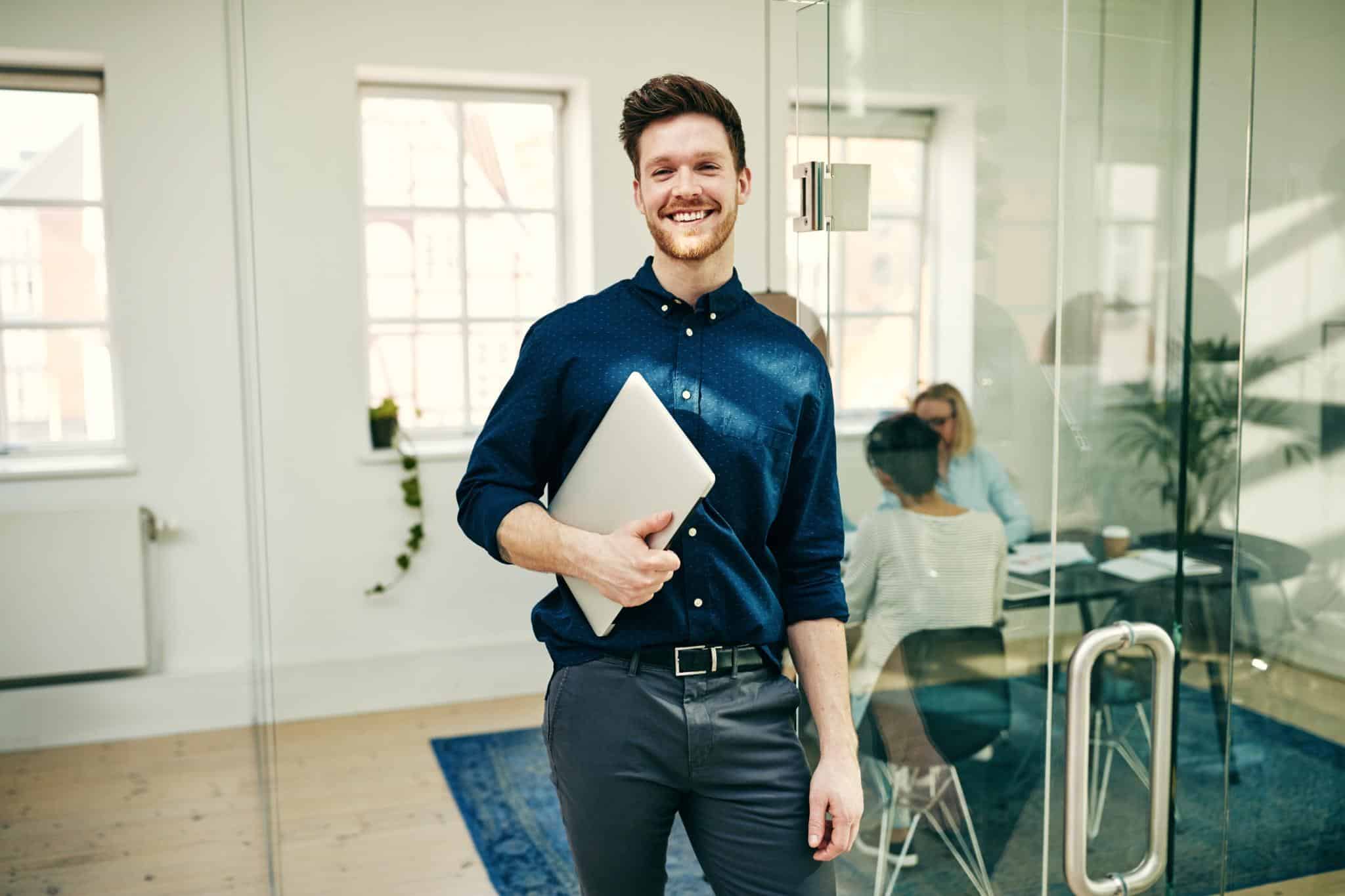 Smiling young businessman holding a laptop in a modern office with glass walls