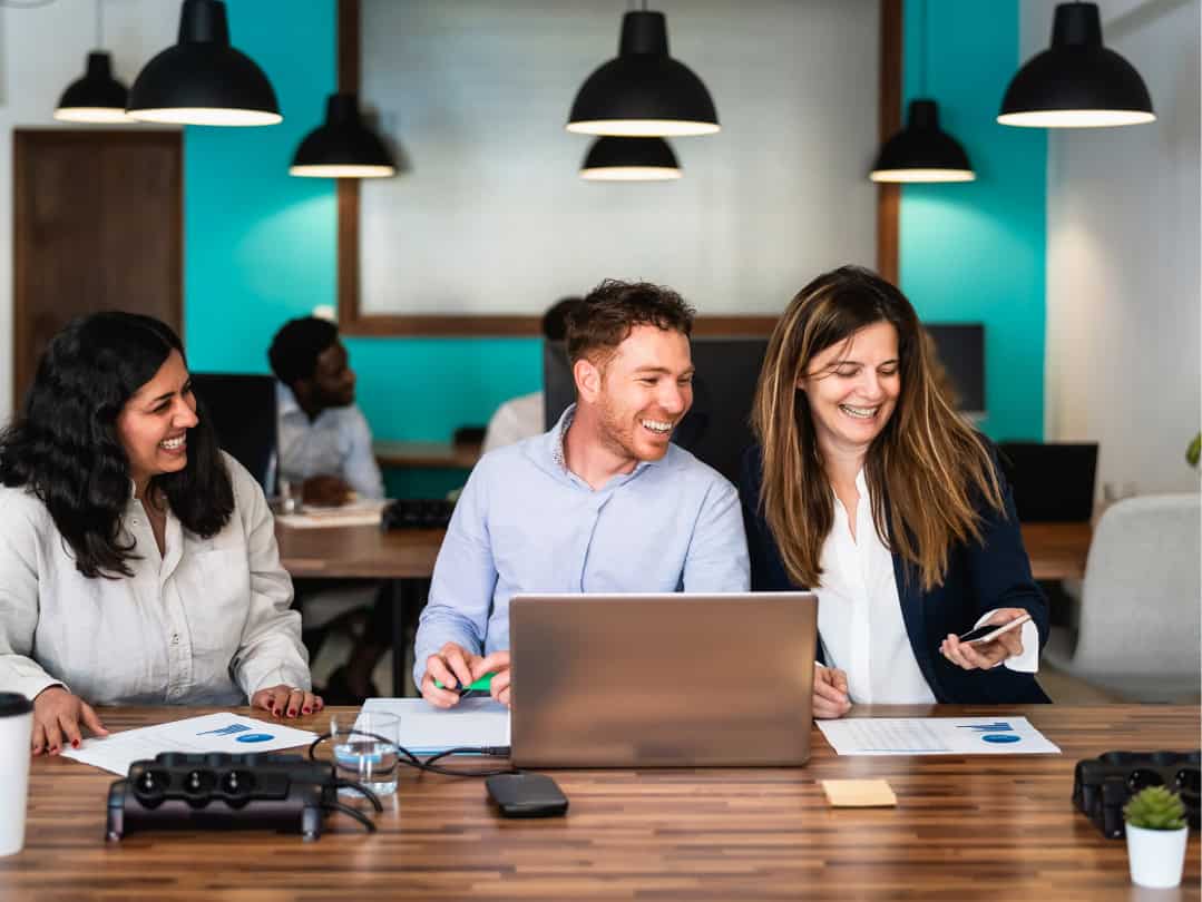 Equipo de trabajo sonriente en una oficina moderna, colaborando en proyectos de marketing digital con documentos y una computadora portátil en la mesa.