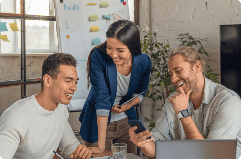 En esta imagen vemos a un equipo de tres personas colaborando en un espacio de trabajo. Parecen estar en una reunión o sesión de lluvia de ideas. Dos hombres y una mujer están reunidos alrededor de una mesa, donde se puede ver un portátil, algunas notas y un vaso de agua. El hombre a la izquierda lleva una camiseta blanca, mientras que el hombre a la derecha tiene un estilo casual con una chaqueta de mezclilla y un smartwatch. La mujer en el centro, que lleva un blazer, está inclinada sobre la mesa, mirando el teléfono que sostiene el hombre a la derecha, mientras sonríe y participa activamente. En el fondo, una pizarra con gráficos y notas adhesivas añade al ambiente de oficina. Esta escena captura un momento de trabajo en equipo, comunicación y un ambiente laboral positivo.