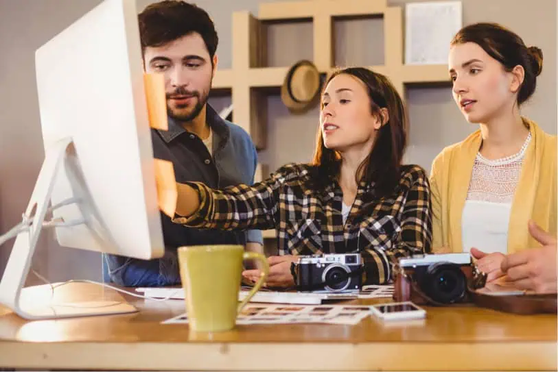 En esta imagen vemos a un equipo de tres personas colaborando en un espacio de trabajo. Parecen estar en una reunión o sesión de lluvia de ideas. Dos hombres y una mujer están reunidos alrededor de una mesa, donde se puede ver un portátil, algunas notas y un vaso de agua. El hombre a la izquierda lleva una camiseta blanca, mientras que el hombre a la derecha tiene un estilo casual con una chaqueta de mezclilla y un smartwatch. La mujer en el centro, que lleva un blazer, está inclinada sobre la mesa, mirando el teléfono que sostiene el hombre a la derecha, mientras sonríe y participa activamente. En el fondo, una pizarra con gráficos y notas adhesivas añade al ambiente de oficina. Esta escena captura un momento de trabajo en equipo, comunicación y un ambiente laboral positivo.