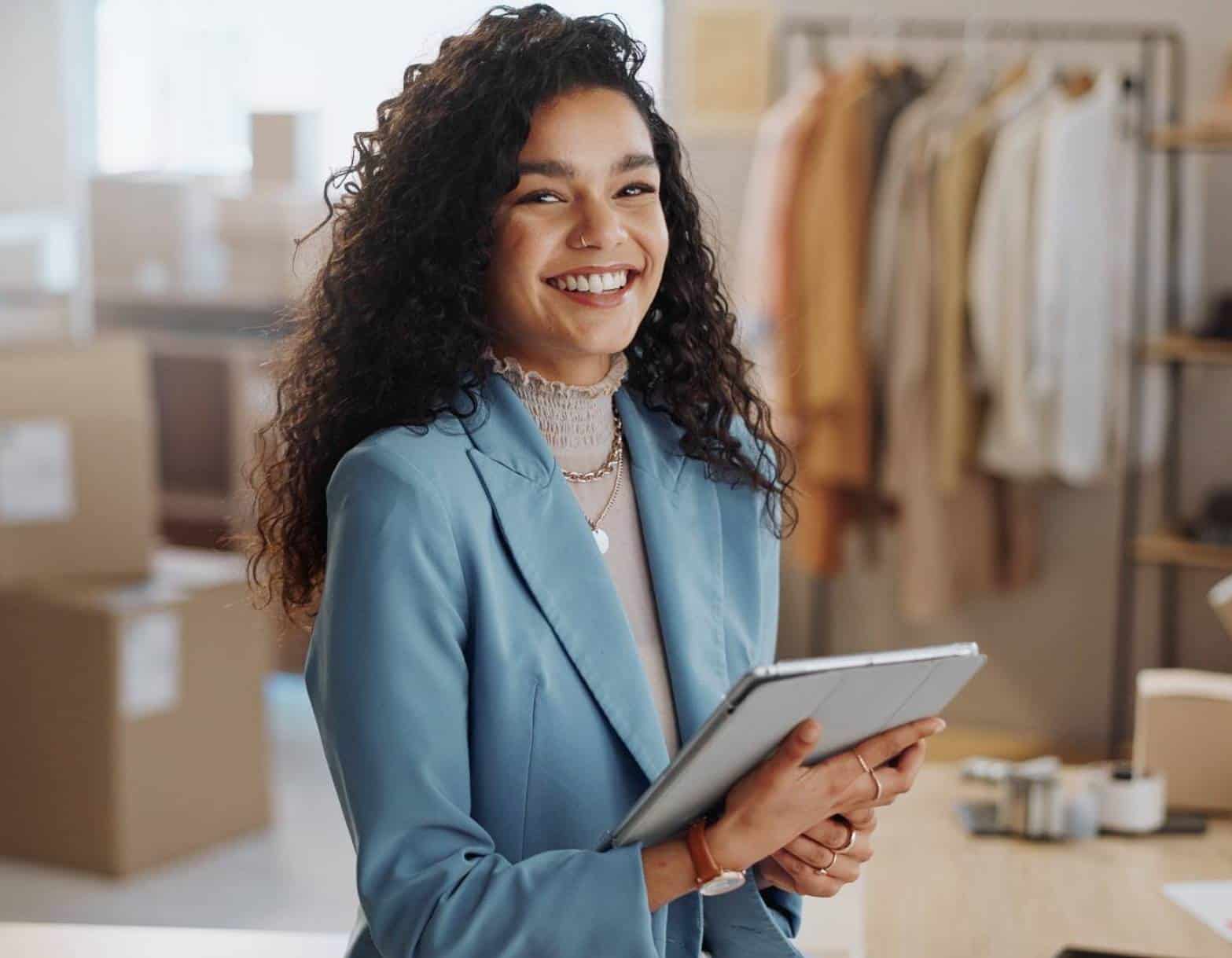 Diseñadora de moda sonriente usando una tablet en su estudio, rodeada de materiales y cajas para su negocio de moda