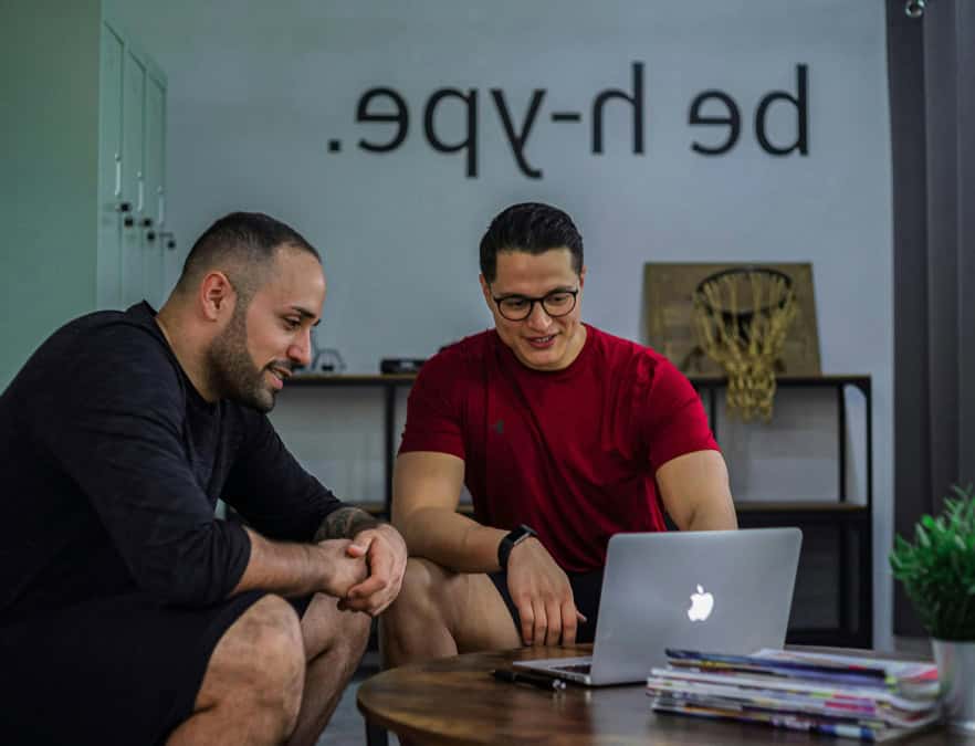 Dos hombres sonrientes mirando una computadora portátil en un entorno informal de oficina o gimnasio, con decoración motivacional en la pared.