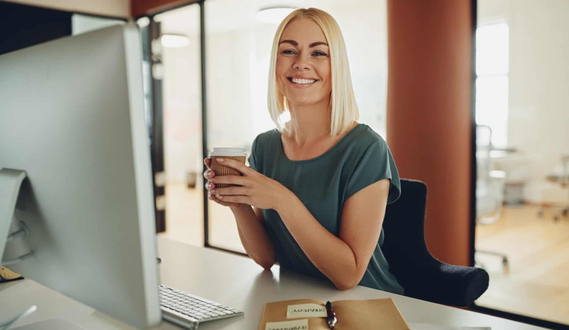 Mujer de negocios sonriente tomando café mientras trabaja en su escritorio en una oficina moderna.