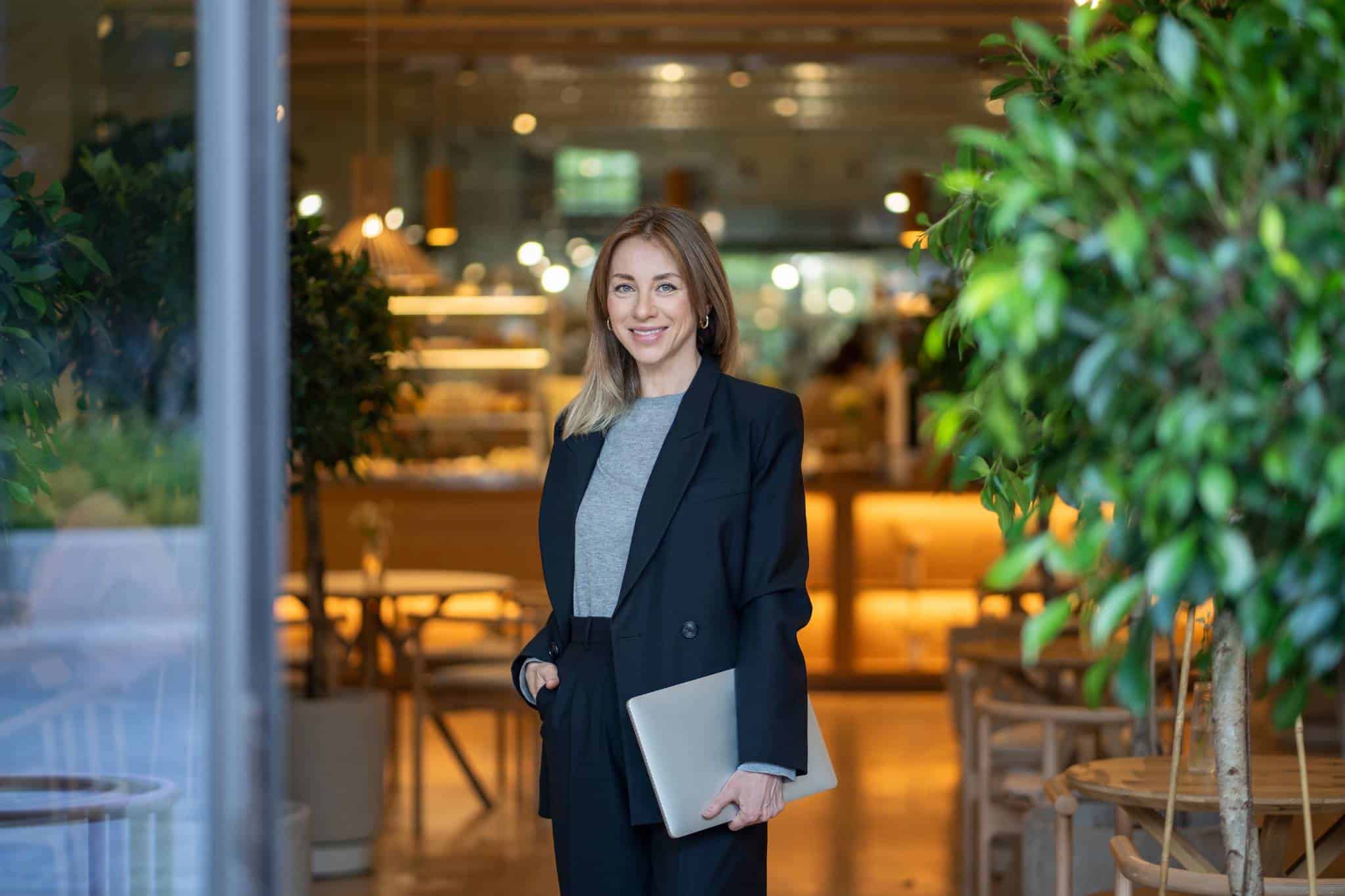 Propietaria de un restaurante posando en la entrada de su negocio con un portátil en mano, lista para gestionar sus operaciones digitales.