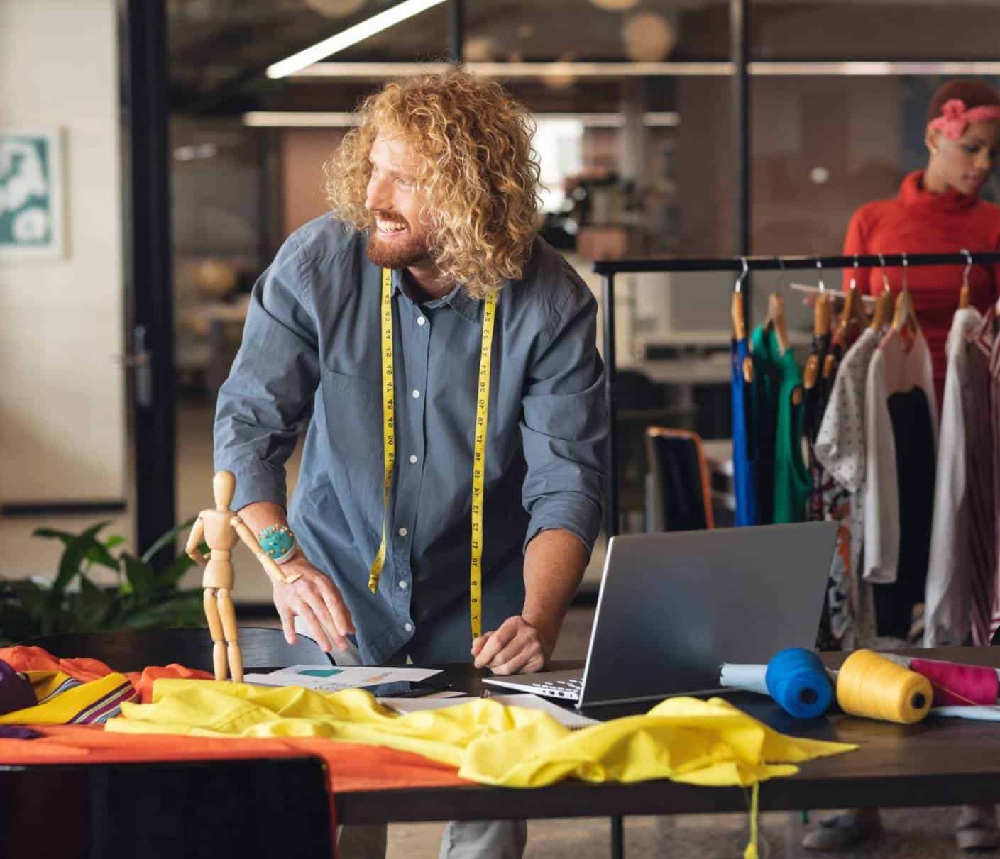 Diseñador de moda sonriente trabajando en su estudio con telas, maniquí y portátil, mostrando creatividad y pasión por el diseño.