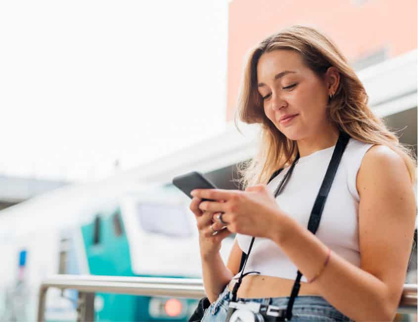 Mujer joven usando su teléfono móvil mientras espera en un entorno urbano, con un tren desenfocado en el fondo.